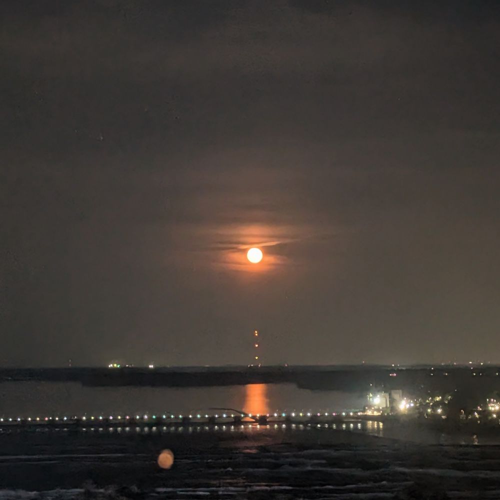 Orange full moon rising above a wide river, reflecting an orange streak on the dark water 
