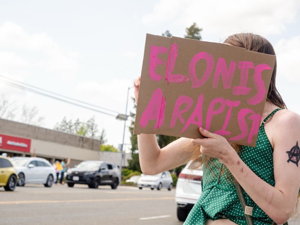 “Elon is a rapist” written on cardboard in pink, held by someone in a green tank top (their face is hidden behind the sign)