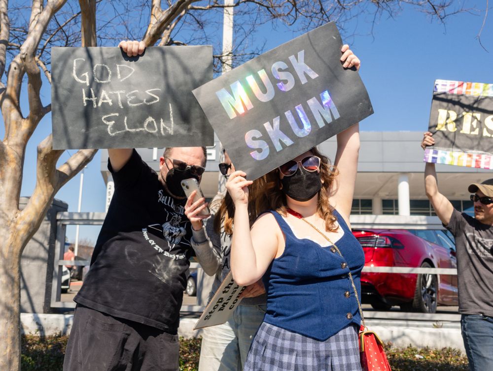 People holding black signs with metallic letters in front of a Tesla dealership: GOD HATES ELON, MUSK SKUM, & RESIST. 