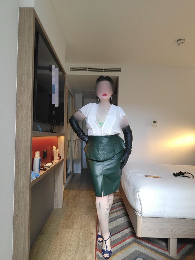 Mistress Jamie stands tall in her navy peep toe high heels, revealing a dark Green polish that matches the leather pencil skirt she is wearing. On the top She wears a lacey Green bra underneath a white, silky, blouse buttoned down slightly to tease and show a hint of her bra underneath. With long black leather gloves to stop on the Upper arm and black slicked back hair with red lips and silver earrings, in the background you can see her various dildos and toys set up for play with a minimal and modern bedroom setting. She invites you to play.