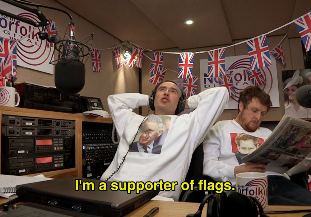 Alan Partridge surrounded by Union Jack banners, wearing a King Charles sweatshirt, relaxing in the North Norfolk Digital offices with Sidekick Simon.