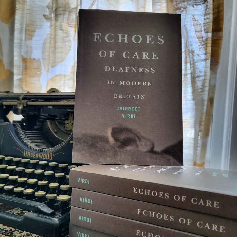 A book standing upright on multiple copies, which are laying next to a 1920s Underwood typewriter.  The book cover is a faded and blurry background, with a photo of a person’s ear horizontal on the bottom, and text emitting from it to represent sound. The text in large font says “Echoes of Care” followed by “Deafness in Modern Britain” and the author’s name, Jaipreet Virdi.