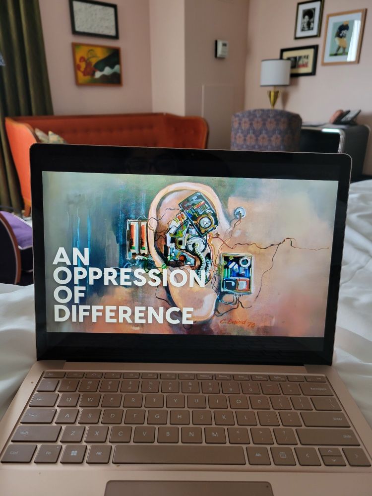 Laptop on a bed with a slide showing  Chuck Baird's art piece of an ear with mechanical parts. Text overlaid says: An Oppression of Difference.
