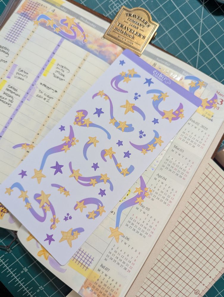 Photo of a sticker sheet of blue and purple stars and clusters of yellow stars with blue to purple gradients. The sticker sheet is sitting on top of a notebook.