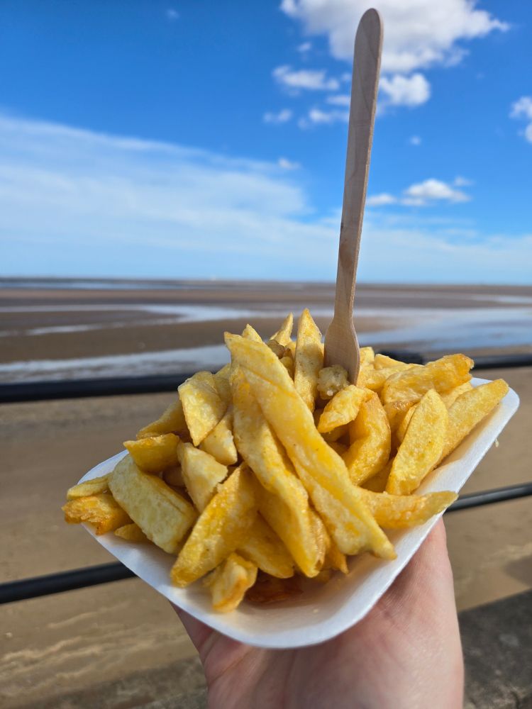 Proper chip shop chips, with a wooden fork, by the beach, under a bright blue sky. 