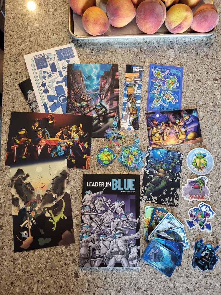 the Full Bundle of the Leader In Blue Zine all laid out on my table. 6 prints, 1 bookmark, 1 sticker sheet, 6 cards, 4 individual stickers, 2 keychains, 1 pin, and the zine itself. all of the above are focused on various iterations of Leonardo from the TMNT.