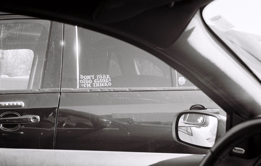 black and white photo of a sticker in a car window. the sticker says “don’t park too close, i’m thick.” the font is a retro/groovy 70’s font with flowers around it. 