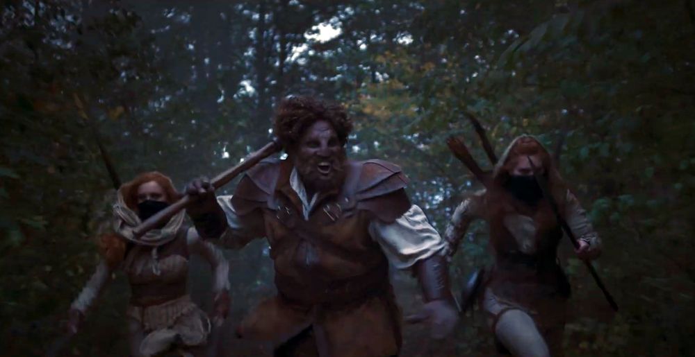 A tense action shot from The Wheel of Time Season 3, showing Bain, Chiad, and Loial running through a dense forest. Bain and Chiad, two Aiel Maidens of the Spear, are dressed in traditional Aiel warrior garb with their faces partially covered, each wielding a spear. Loial, the Ogier, charges forward alongside them, wearing leather armor and carrying a large weapon over his shoulder, his expression fierce and determined. The background is blurred with trees and foliage, emphasizing their movement and urgency.