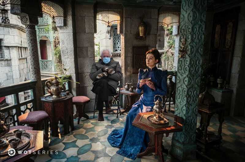 Moiraine Sedai from The Wheel of Time sits elegantly in a lush, intricately designed sitting area, sipping tea in her iconic blue dress. Next to her, Senator Bernie Sanders, photoshopped in his famous mittens and coat from the 2021 inauguration, sits casually on a stool, adding a humorous juxtaposition to the serene setting