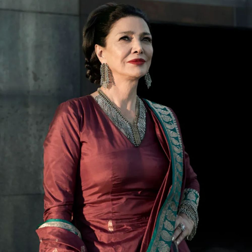 Actor Shohreh Aghdashloo in a still from The Exapnse, stands confidently wearing a richly colored maroon gown with intricate green and gold embroidery. Her hair is styled in an elegant updo, and she wears bold chandelier earrings. The background features a dark, modern architectural setting, emphasizing her poised and commanding presence.