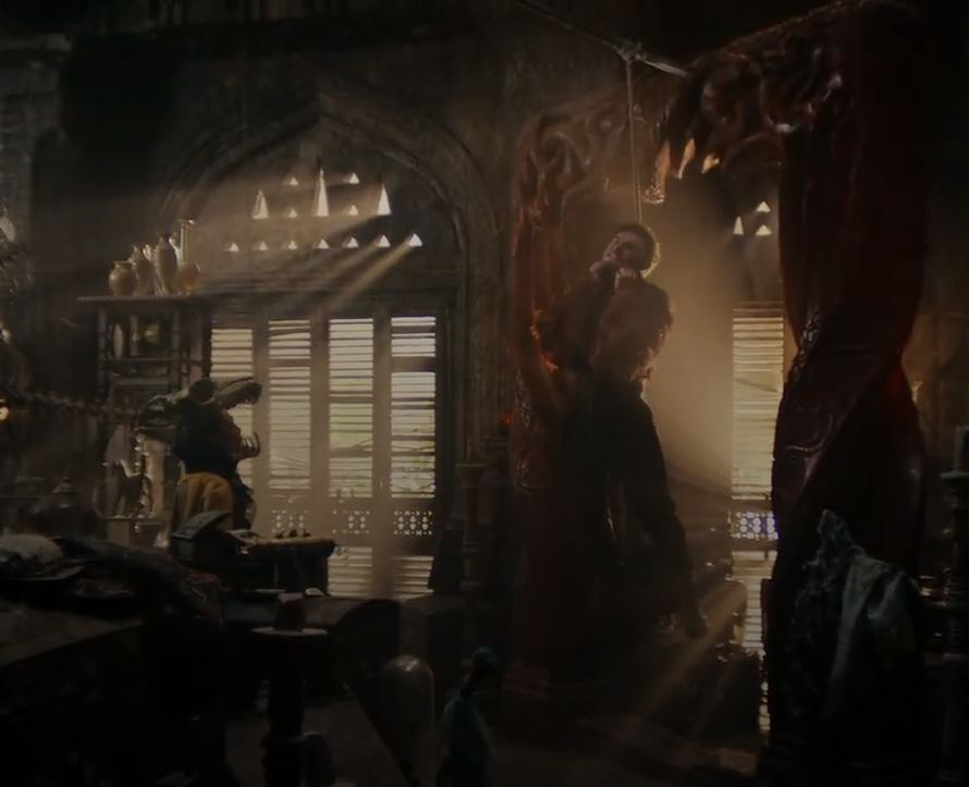 Mat Cauthon hangs limp by the neck from a noose in a dimly lit, ornate chamber, illuminated by shafts of light streaming through shuttered windows. He is suspended just outside the carved red door ter’angreal. The scene is filled with shadow, mystery, and a haunting stillness. Chesmal emery watches on. The rope seems to be held up by a spear driven into the red door frame