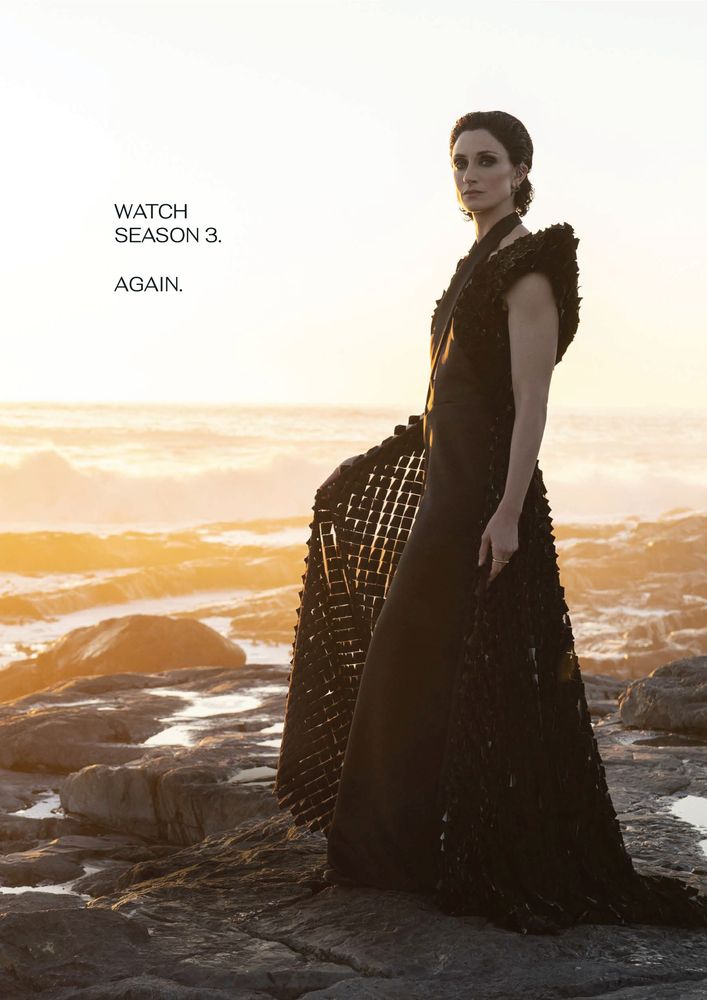 Natasha O'Keeffe as Lanfear from The Wheel of Time, standing on rocky terrain by the ocean at sunset. She wears a dramatic black gown with a structured, textured design featuring a grid-like, flowing outer layer. Her dark hair is slicked back, and she gazes confidently into the distance, illuminated by the golden light of the setting sun behind her, with waves crashing in the background. The caption 'Watch Season 3. Again.' appears to the left of the image. 