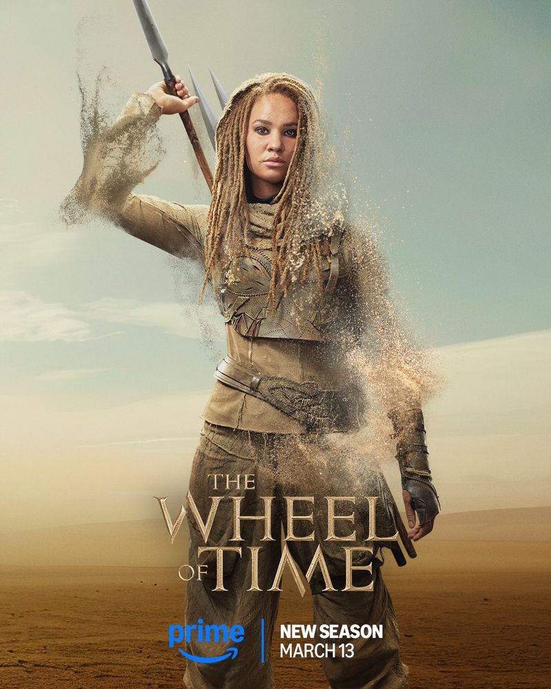 A promotional poster for The Wheel of Time Season 3 features Aviendha, played by Ayoola Smart, standing in a vast desert landscape. She is dressed in Aiel warrior garb, with layered earth-toned clothing and leather armor. She grips a pair of spears, one raised above her shoulder in a battle-ready stance. The right side of her body is dissolving into sand, blending into the arid background. The show’s title, The Wheel of Time, appears in gold at the bottom, with the Prime Video logo and the release date, March 13, beneath it.
