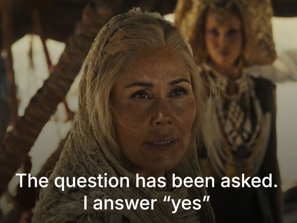 Nukâka Coster-Waldau as Bair, wearing desert-toned Aiel clothing with a hooded shawl, standing inside a tent with a determined expression. The subtitle reads: “The question has been asked. I answer ‘yes.’”