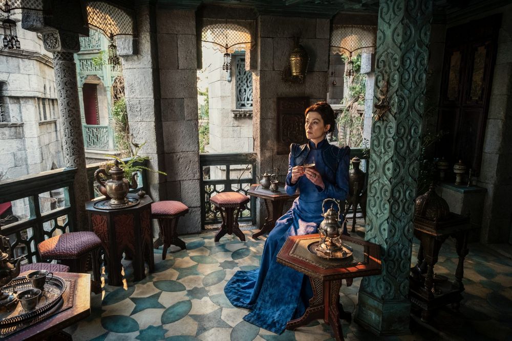 Rosamund Pike as Moiraine sits alone in an ornately decorated terrace, dressed in a regal blue gown, holding a small cup and looking into the distance. The setting features intricate stonework, patterned floors, and elegant furnishings.
