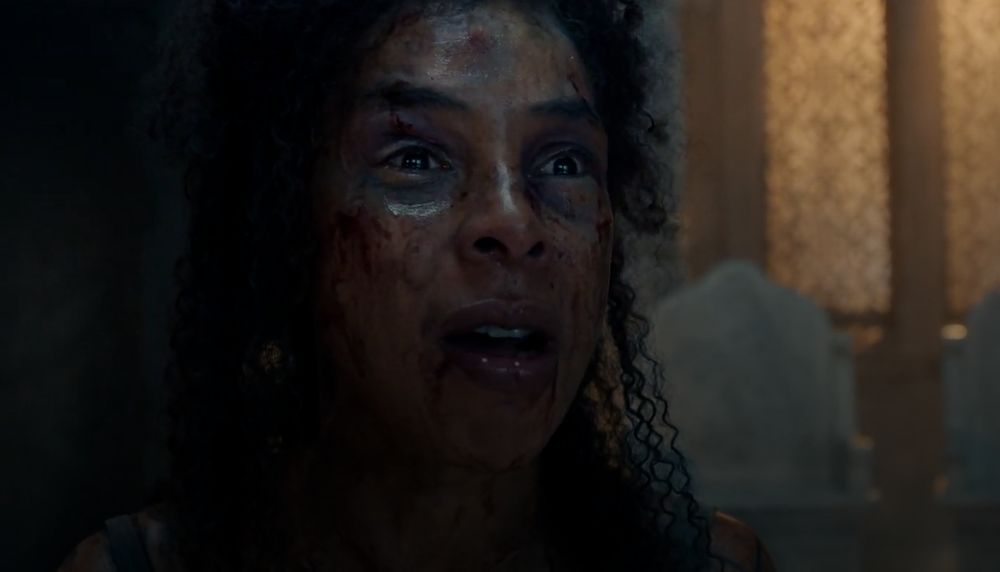 Sophie Okonedo as Siuan Sanche in The Wheel of Time episode 308, her face bloodied and bruised, eyes wide with anguish and emotion. She is in a dimly lit hall with ornate, high-backed chairs visible in the background.