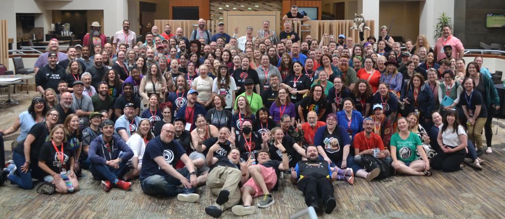 A large group photo of enthusiastic Wheel of Time fans gathered at WoTCon, an annual fantasy convention held in Columbus, Ohio. Attendees of diverse ages and backgrounds smile, pose, and celebrate their shared love of the series. Many wear themed shirts, lanyards, and costumes. The atmosphere is joyful and welcoming, capturing the strong sense of community and excitement among over 200 fans united by their passion for The Wheel of Time.