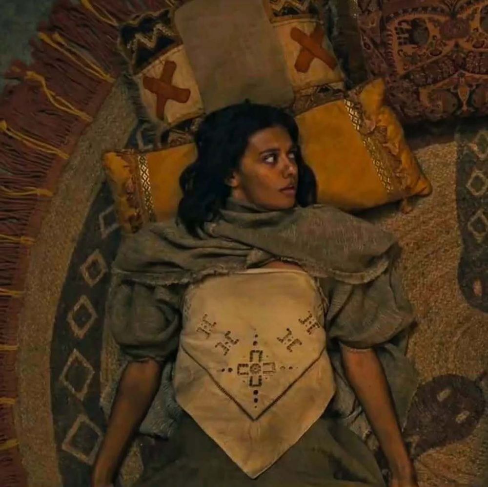Madeleine Madden as Egwene al'Vere from the Wheel of Time Season 3 teaser trailer lies on a woven rug, surrounded by colorful pillows. She appears weary, wearing layered clothing with a shawl draped over her shoulders and an embroidered apron-like garment on her chest. Her expression is serious, and her gaze is turned to the side, as if reacting to something out of frame.
