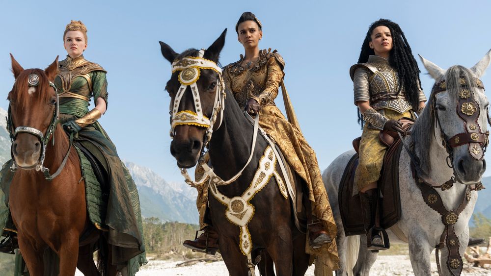 The image features Ceara Coveney as Elayne Trakand, Madeleine Madden as Egwene al’Vere, and Zoë Robins as Nynaeve al’Meara from The Wheel of Time Season 3. The three women are mounted on horses, exuding strength and determination as they prepare for an important journey or confrontation. Elayne, on the left, is clad in a regal green gown with gold accents, befitting her royal status. Egwene, in the center, wears an intricate golden outfit adorned with ornate detailing, reflecting her growing power and authority. Nynaeve, on the right, is armored in silver and gold, conveying her readiness for battle. Each horse is elaborately adorned with matching bridles and saddles, adding to the grandeur of the scene. Set against a backdrop of clear skies and mountainous terrain