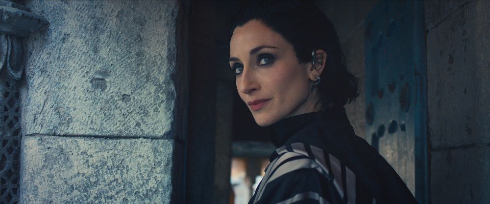 The image features Natasha O’Keeffe as Lanfear, one of the Forsaken, in The Wheel of Time Season 3. Released by Prime Video as part of a first-look series, the photo captures Lanfear’s enigmatic presence. She is shown in a stone-walled setting, her expression calm yet piercing, with a subtle smirk that hints at her cunning and confidence. Her dark, sleek hair is styled back, and she wears elegant, striped attire that reflects her powerful and mysterious character.