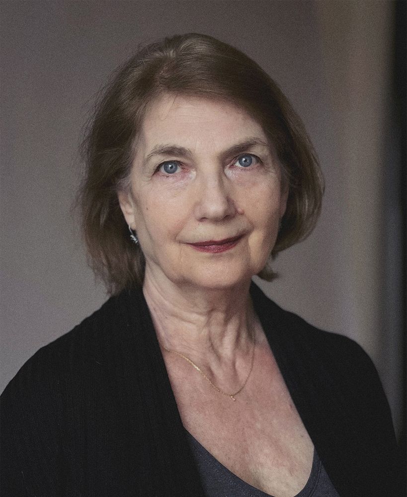 Headshot of actor Zuzana Hodkova, who has been cast to play Lini in The Wheel of Time. The image shows her looking directly at the camera with a calm and poised expression. She has short, light brown hair and is wearing a simple black cardigan over a gray top, accessorized with a delicate gold necklace and earrings. The background is softly blurred, emphasizing her thoughtful demeanor.