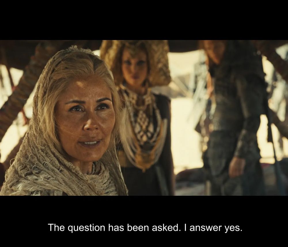 Nukâka portrays Bair in The Wheel of Time Season 3, Episode 4. She stands in a sunlit Aiel tent, wrapped in traditional desert-colored shawls, her expression calm yet resolute. Behind her, Sevanna and Couladin. The warm, earthy tones of the scene emphasize the harsh desert setting. The subtitle reads, “The question has been asked. I answer yes”