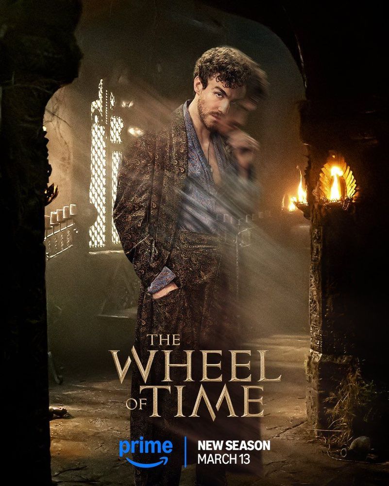 A promotional poster for The Wheel of Time features Dónal Finn as Mat Cauthon, standing in a dimly lit, ancient corridor with intricate stonework and flickering torches mounted on the walls. He wears an ornate, patterned robe over a loose blue shirt, with his hand raised to his face in a mysterious, knowing gesture. His expression is both mischievous and intense, and a blurred motion effect makes it appear is though he is separating from his own body. The show's title, The Wheel of Time, is displayed prominently at the bottom in gold lettering, with the Prime Video logo and the text "New Season March 13" beneath it.

