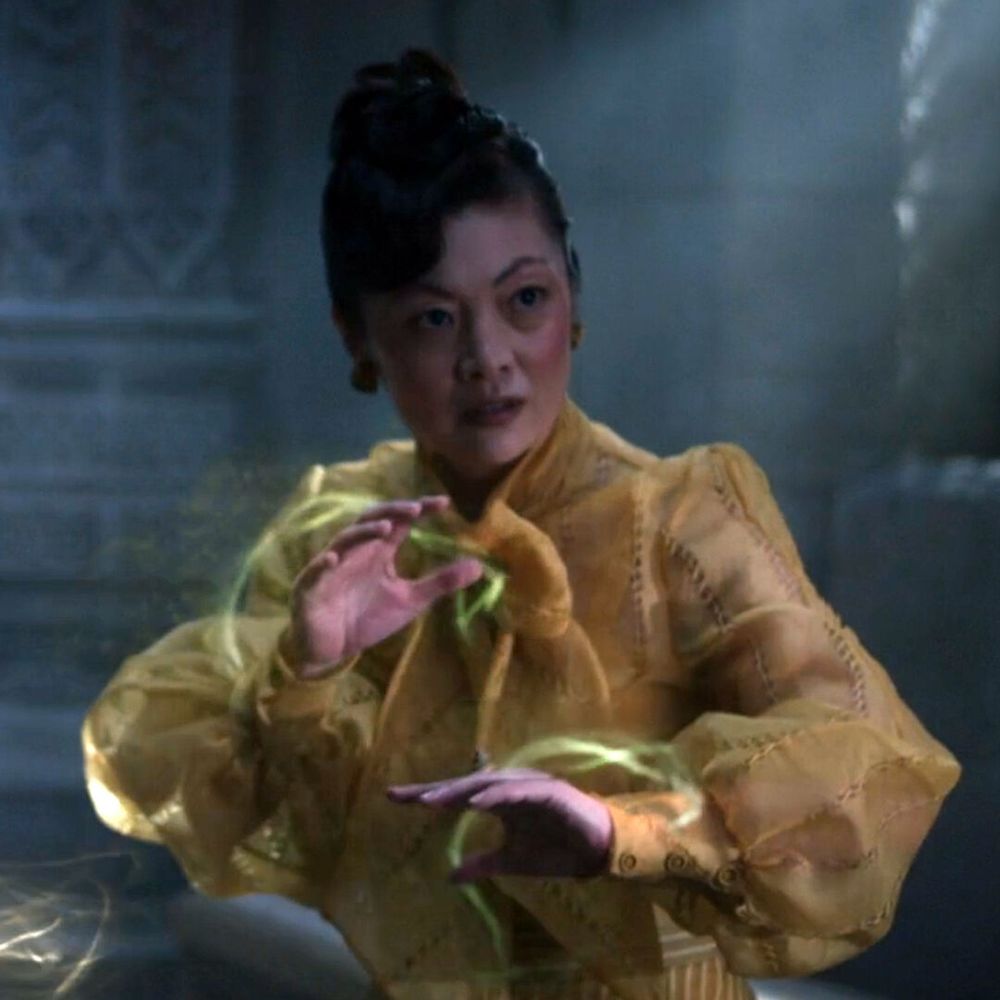 A dimly lit scene from The Wheel of Time features an unknown actress portraying Chesmal Emry, a member of the Black Ajah. She wears a richly detailed yellow blouse with puffed sleeves and intricate embroidery. Her dark hair is styled in an elegant updo, and she has an intense, focused expression. Greenish-yellow weaves of the One Power swirl around her hands, illuminating the air with a faint magical glow. The background is shadowy and atmospheric, with hints of ornate stone architecture visible through the misty lighting.