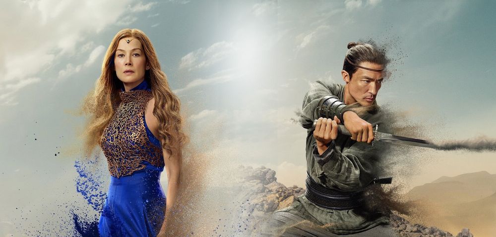 combined image from The Wheel of Time Season 3 posters featuring Moiraine Damodred (played by Rosamund Pike) and Lan Mandragoran (played by Daniel Henney). Moiraine stands on the left, wearing a regal blue gown with an intricate gold lattice bodice, her long hair flowing as she gazes forward with a determined expression. Blue energy and sand swirl around her, dissolving into the background. On the right, Lan is poised in a battle stance, gripping his sword with both hands. He wears green and gray warrior attire, a leather belt, and bracers. His expression is focused and intense, with the right side of his body and sword fragmenting into sand. 