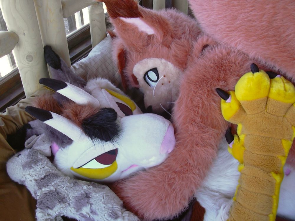 A closeup of two fursuits cuddling. Pipsqueak the squirrel is laid on top of Vec the dragon, their arm wrapped around Vec’s neck, and Vec’s paw resting on Pipsqueak’s side. Bunny the stuffed plush rabbit lays on the pillow next to Vec’s head. 