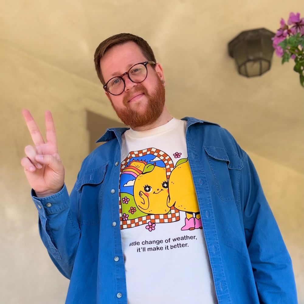 Picture of me wearing a blue over shirt and a graphic tee. My head is cocked to the right and I’m giving a peace sign. The graphic tee depicts a lemon wearing pink boots looking into a mirror. The mirror has an orange and yellow checkerboard border. In the mirror behind the lemon there is a sunshine with a smile on its face peeking out from behind a rainbow and below is a field of flowers. Text below the image reads “little change of weather, it’ll make it better”.