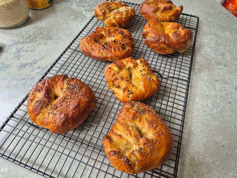 7 mishappen bagels on a cooling rack. Assorted toppings. 