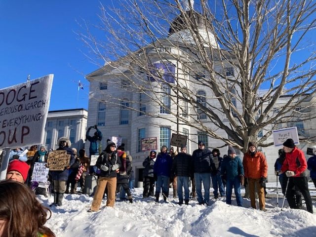 A crowd of protestors bundled up on a cold winter day for the 50501 rally at the Maine State House in Augusta. In the for round is a drift of snow, with dozens of people facing the camera behind it. Behind them is a huge leafless tree and behind that is the back of the State House building against a clear blue sky
