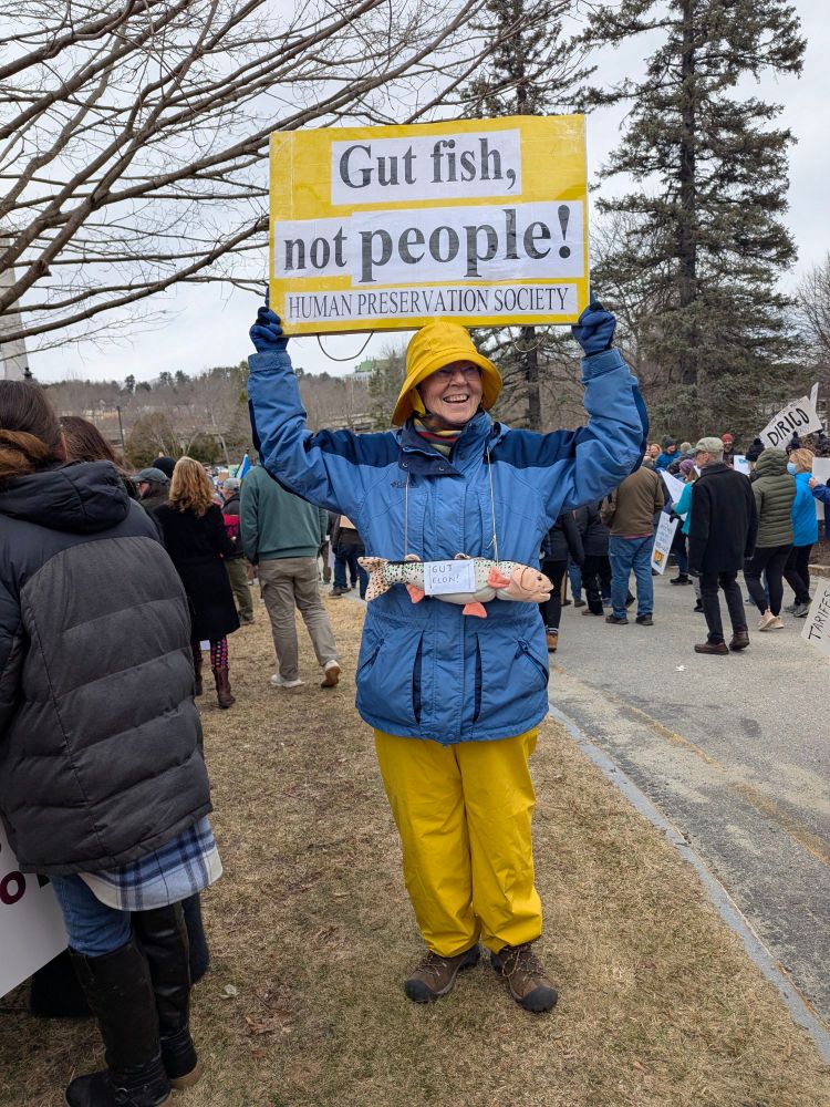 A smiling woman dressed as a fisherman/person in yellow rain pants and hat with a blue jacket holds up a sign "Gut fish, not people! Human Preservation Society" To complete the ensemble, she has a stuffed fish on a cord hanging from her neck.