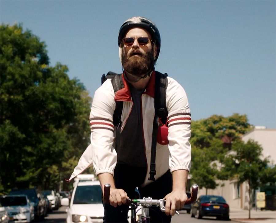 O cara de High Maintenance andando de bicicleta com mochila, óculos escuros e capacete. na mochila tem uma caixa de som pendurada.