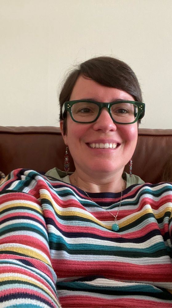 Image of a smiling, brunette woman with dark green glasses, short hair and a story jumper. 