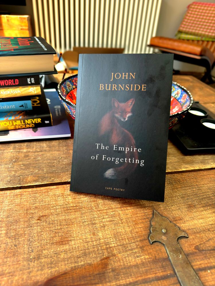 A thin, paperback book of poetry propped up on a table. The book has a dark cover, with a shadowy fox turning its head to look back at the reader. Other books, out of focus, can be seen behind it. 