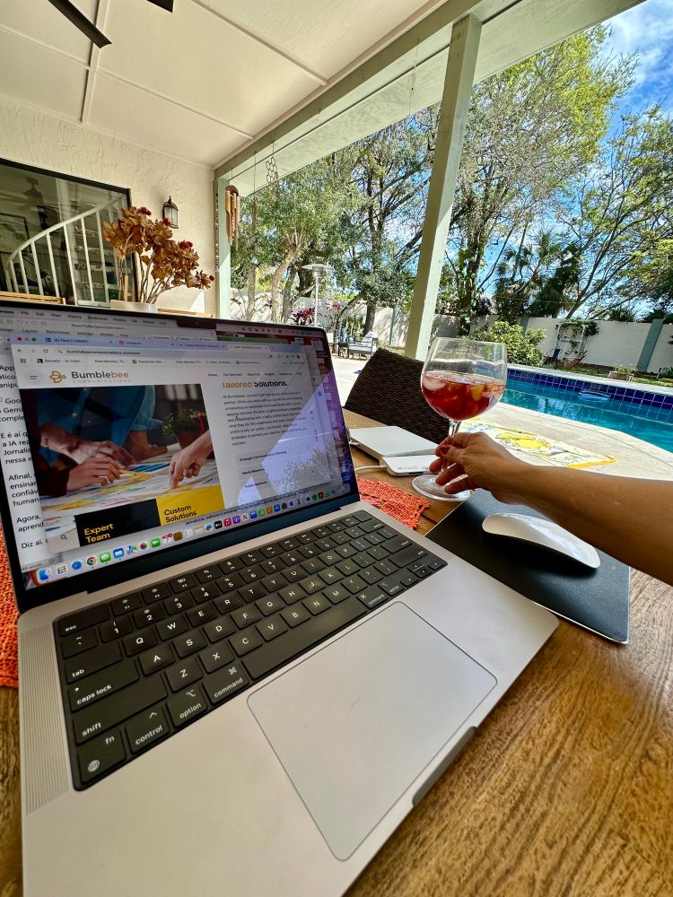 The photo shows a cozy backyard setting with a wooden table. On the table, there’s a laptop open, displaying a webpage with colorful images and text. Next to the laptop, a female hand holds a glass of red drink, a red wine sangria, with a few pieces of fruit visible inside. In the background, there is a swimming pool surrounded by greenery, and sunlight filters through the trees, creating a warm and inviting atmosphere on this Friday afternoon. The scene conveys a sense of relaxation and enjoyment, as we celebrate Women’s International Day! 