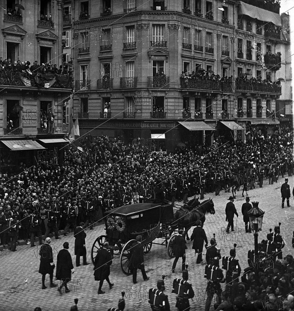 Black and white photo of a huge crowd lining the streets and windows and balconies of a Paris street watching a black carriage drawn by black horses and escorted by a group of men in black suits passes on a cobblestone street