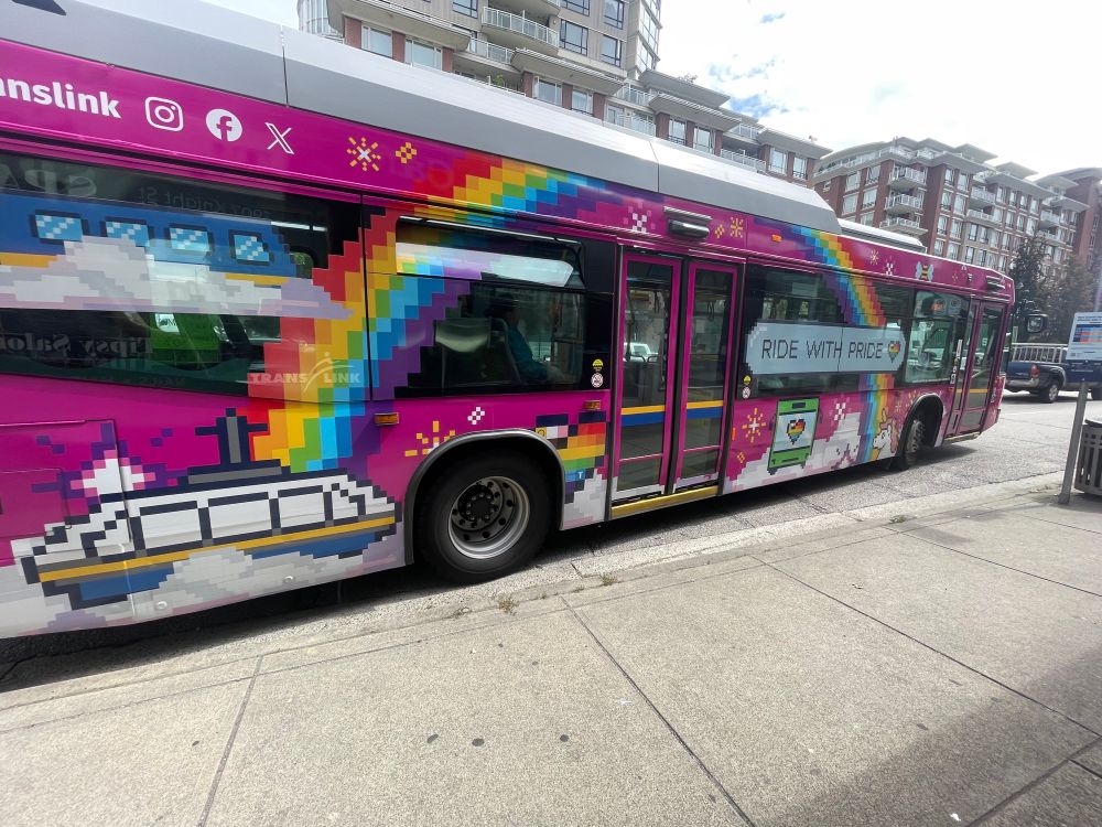 Vancouver’s pride bus, side shot:
The bus has a pink 8-bit-style wrap with a large, pixelated rainbow arcing over the last 2/3 of the bus. There’s a white unicorn with an orange mane, and a bus with a rainbow heart on the bottom-centre panel of the bus with a cloud. The back panel has a tram and the base of the rainbow. There is a large, light blue text bubble that says “RIDE WITH PRIDE” with a rainbow heart in the direct centre of the side of the bus. 