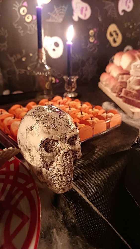 Imagem mostrando a decoração de uma festa de halloween. Uma caveira na frente e docinhos laranja atras, com velas pretas. 