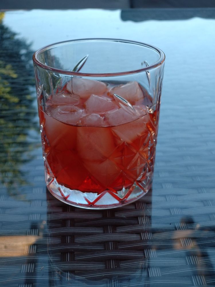 An Old Fashioned glass filled with ice cubes and an day-glo orange coloured drink that is the Negroni. It sits on a glass topped outside table.  