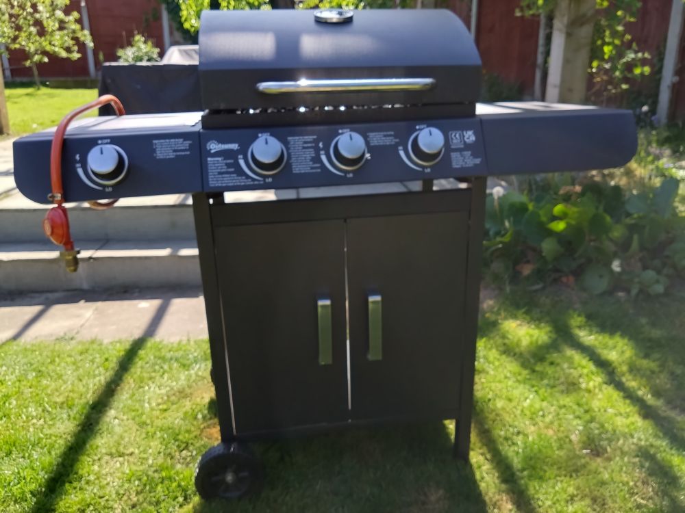 A gas fired barbecue sits menacingly in a garden. Three burner controls and a ring control. At the bottom are two cupboard type doors. Why it has two doors is beyond me as the sides and rear are open. A gas pipe and regulator dangle provocatively, enticing immediate urgent connection. 