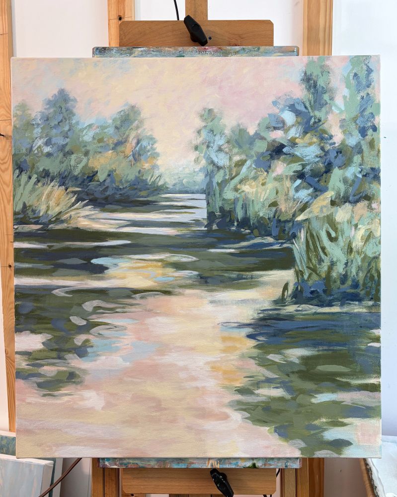 A painting held on an easel depicting a river in neutral pinks and dark greens flowing into the distance where there is a warm light in the sky with trees and foliage along the bank and light and dark reflections in the water. 