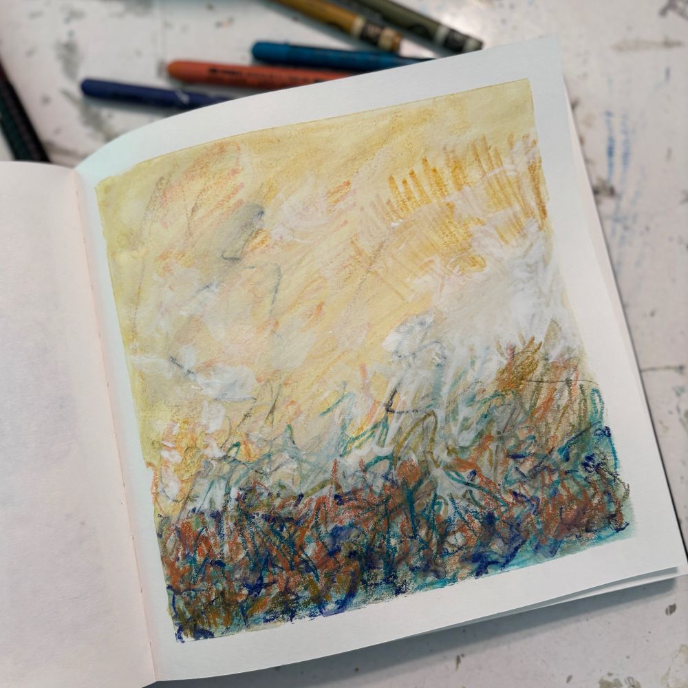 A sketchbook open on a table showing a mixed media drawing with expressive calligraphic marks in blue-greens and orange on the bottom third of the page over soft yellows covering the top two thirds.