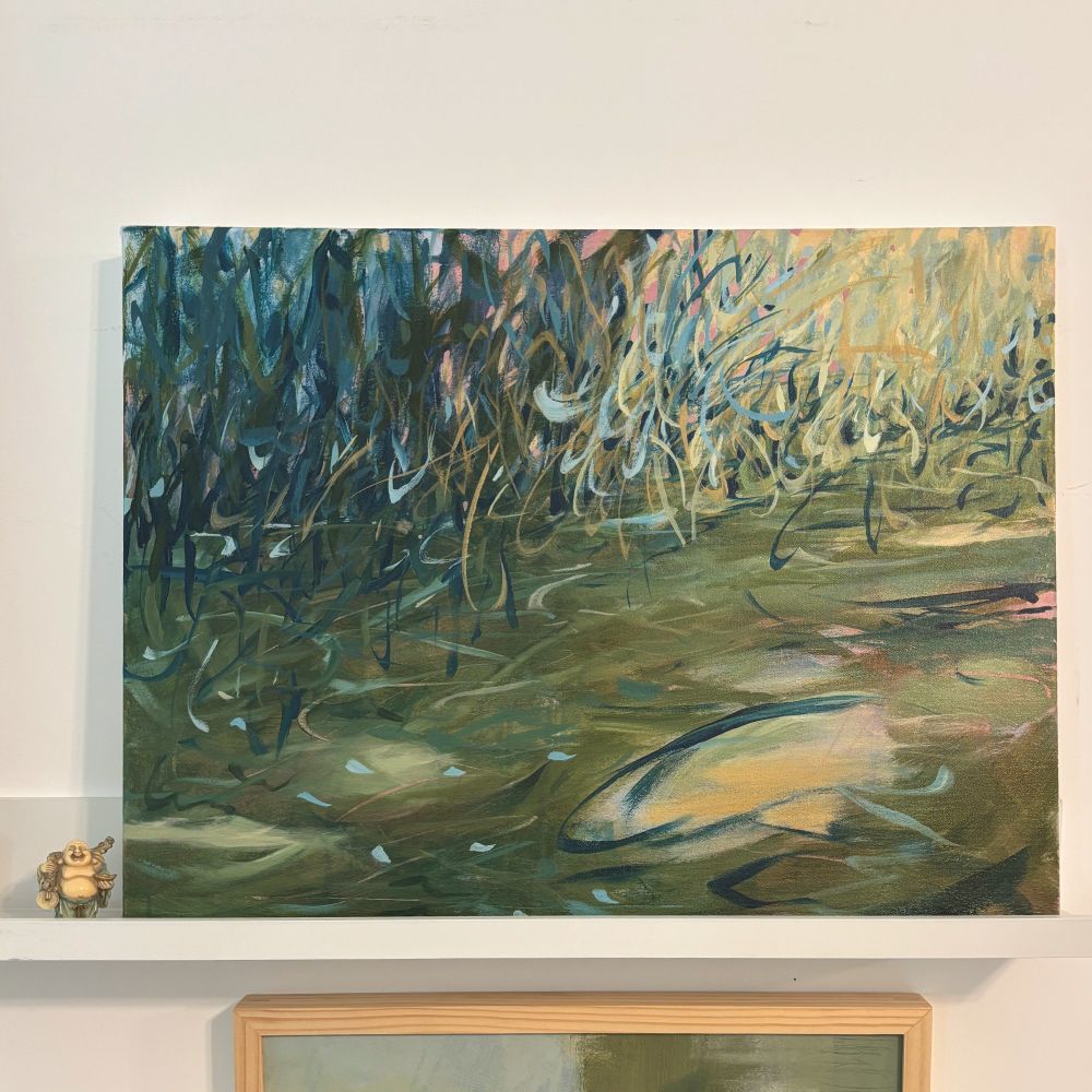 A painting on canvas sitting on a shelf next to a small Buddha figurine. This abstract painting suggests a water's edge with expressive marks in blue and green on a soft green surface with light yellow highlights suggesting filtered light on water.