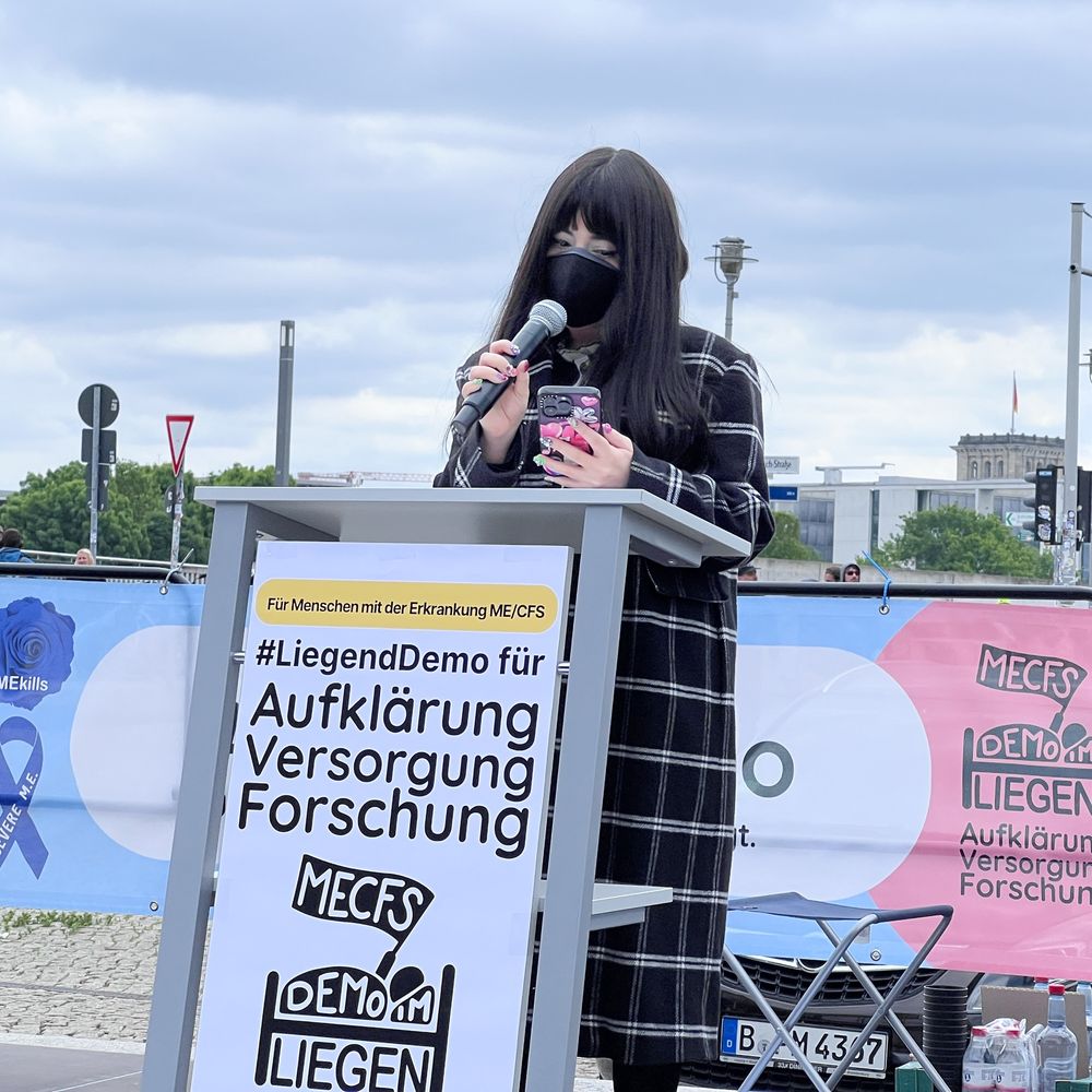 A young woman with long dark hair stands at a podium with a microphone, wearing a black face mask and checkered coat. The podium sign reads: “#LiegendDemo für Aufklärung, Versorgung, Forschung – Für Menschen mit der Erkrankung ME/CFS”. Banners and protesters can be seen in the background.