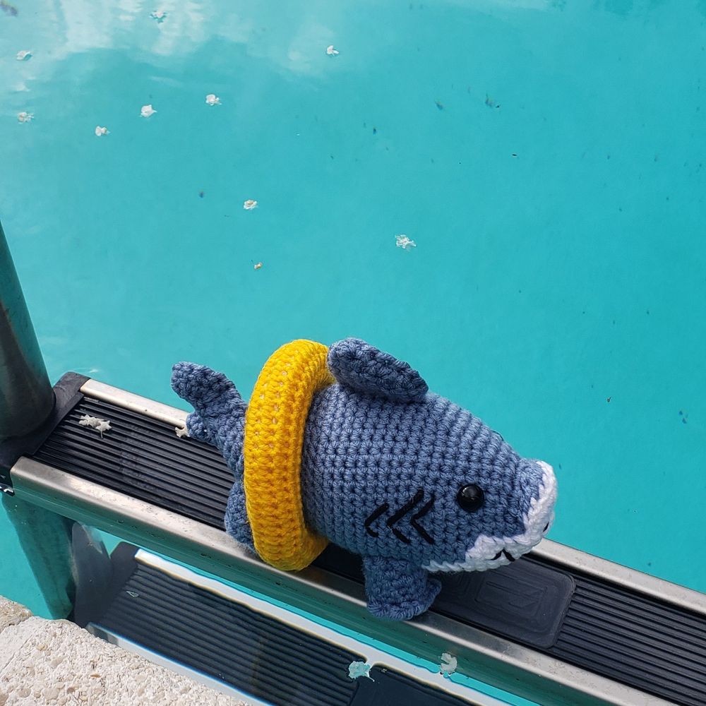 A crochet shark wears a yellow safety floatie. The shark also has legs. It sits on the top step of a pool ladder, near the teal-colored water.