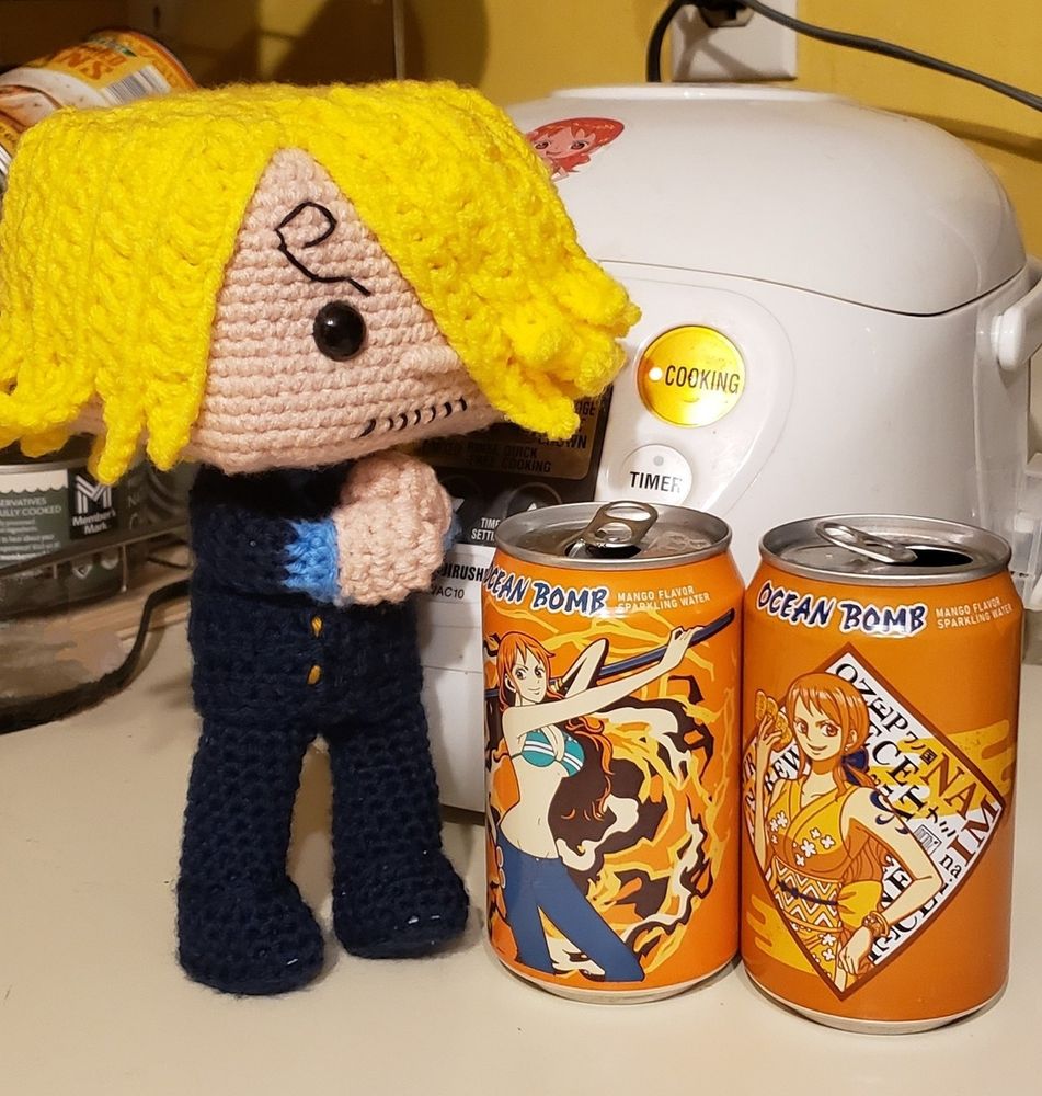 An amigurumi (crochet) Sanji Blackleg stands on a countertop with his hands on his heart. He's looking at two Ocean Bomb cans with Nami on them. If a crochet guy's nose could bleed, his would be.