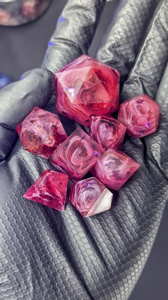 You see a black gloved hand holding a bunch of dice blanks. They're made of warm pink/red tones with orange glitter and purple flakes. Inside each die is a swirl of opaque lilac, making it look like spirals within a rose.